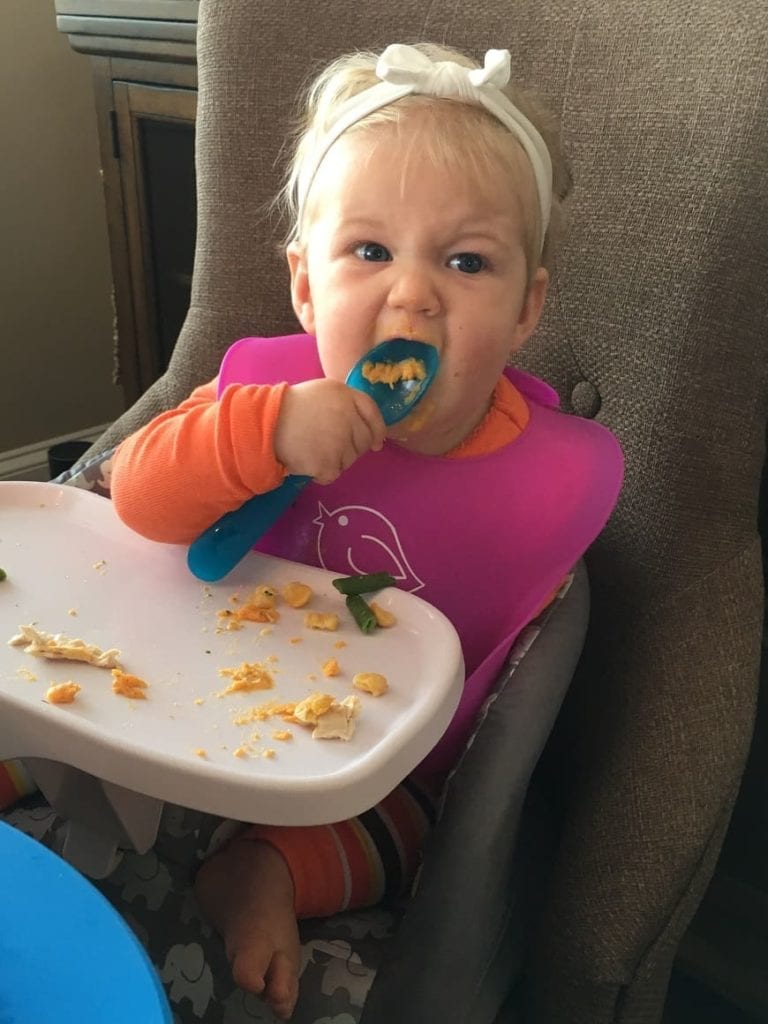 7 month baby holding blue spoon in high chair eating food through baby led weaning