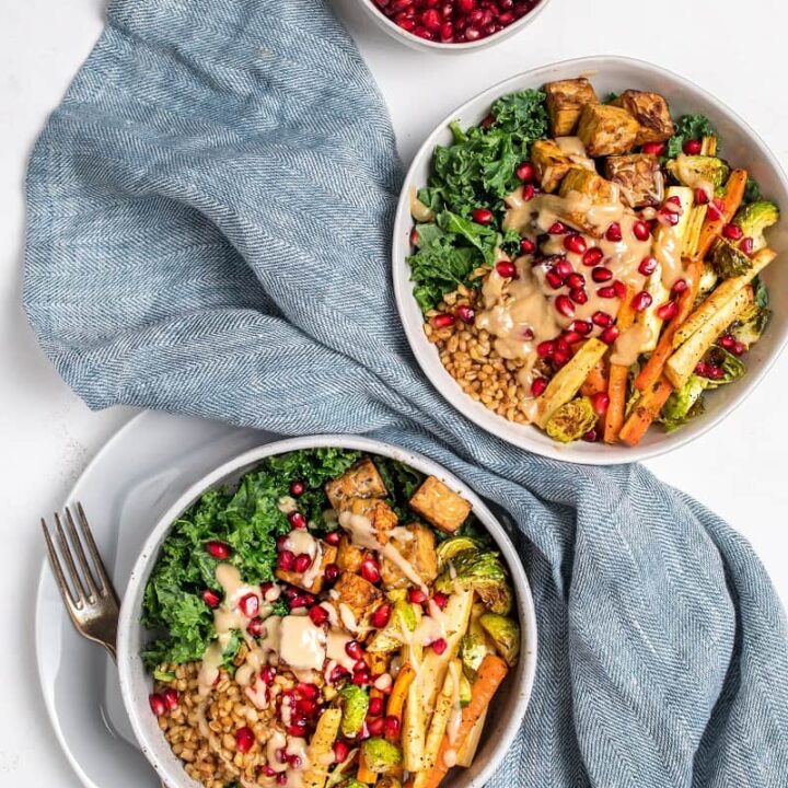 two white bowls with roasted vegetables and topped with pomegranate seeds and tahini dressing next to blue counter cloth