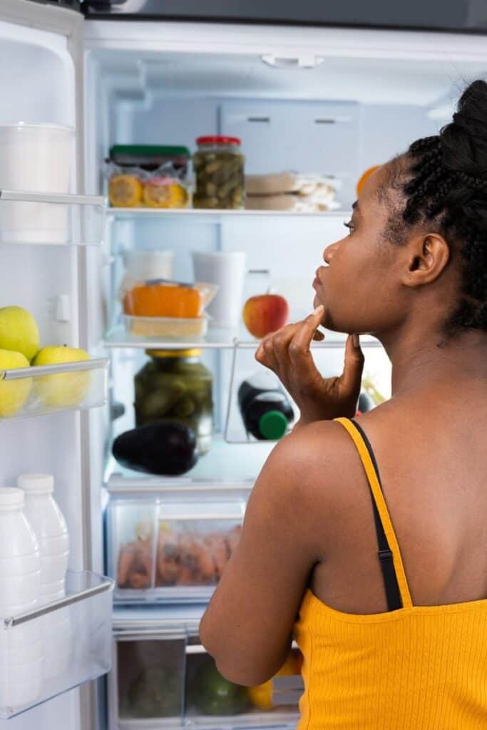 woman in yellow tank top looking into open refrigerator to decide what to eat