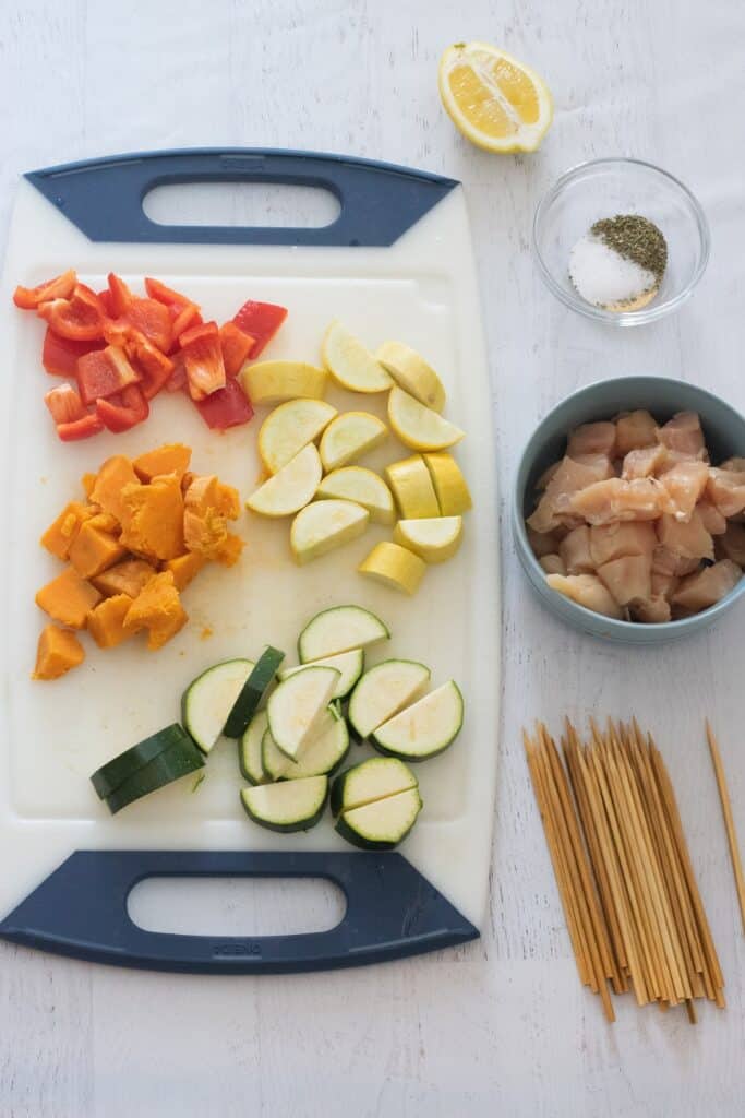 chopped vegetables on white cutting board next to blue bowl with raw chicken to make oven baked chicken kebabs