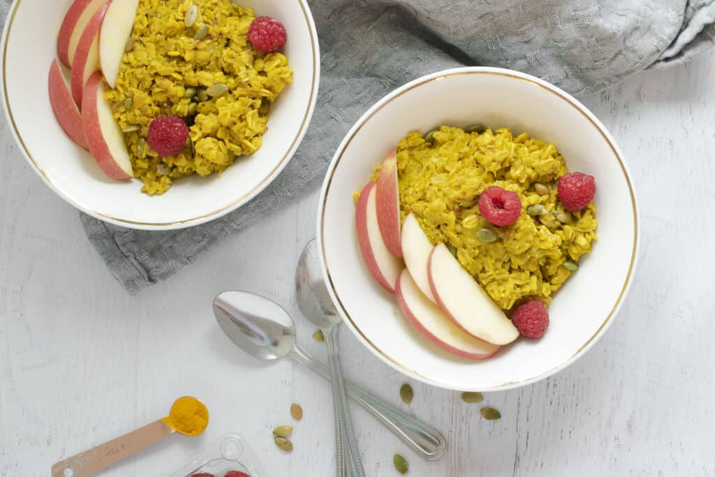 turmeric oatmeal in white bowls topped with raspberries, apple slices and pumpkin seeds