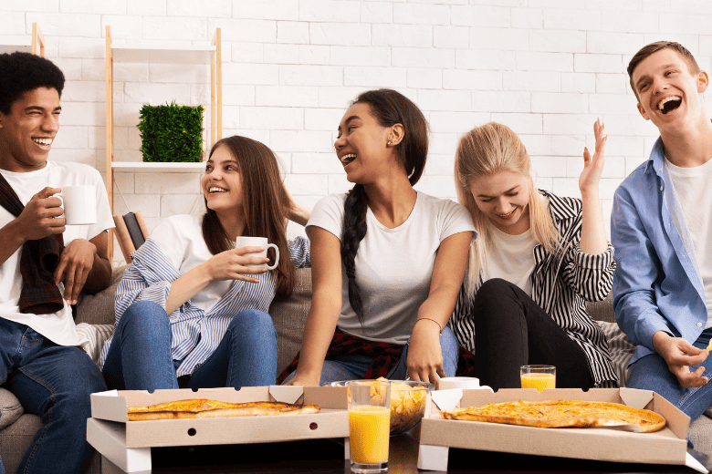 teenage kids laughing on couch with boxes of pizza