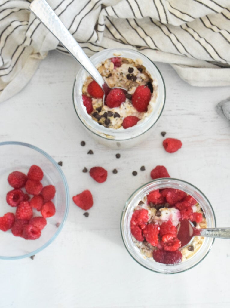 glass jars with overnight oats with raspberries and yogurt with extra raspberries and chocolate chips scattered on the table