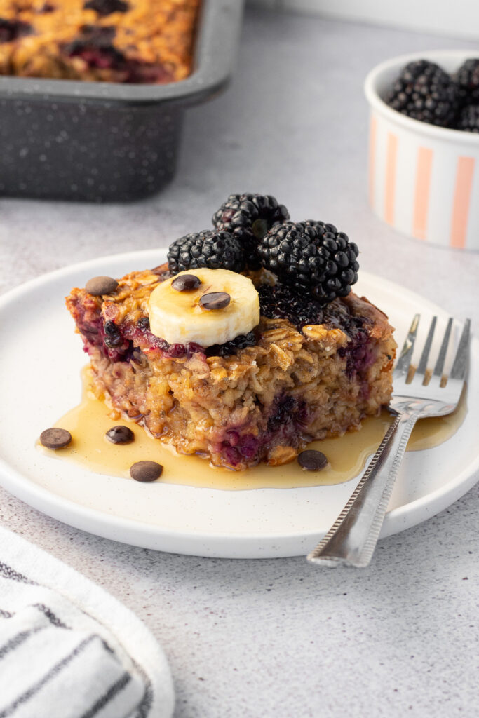 Maple blackberry baked oatmeal topped with fresh blackberries and banana slices, served on a white plate with a fork.