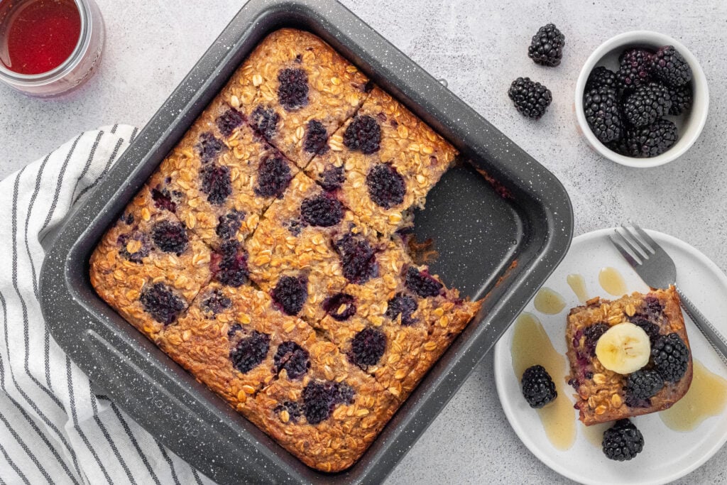 This image features a freshly baked maple blackberry oatmeal in a baking dish, served with a slice on a plate topped with banana slices, surrounded by fresh blackberries.